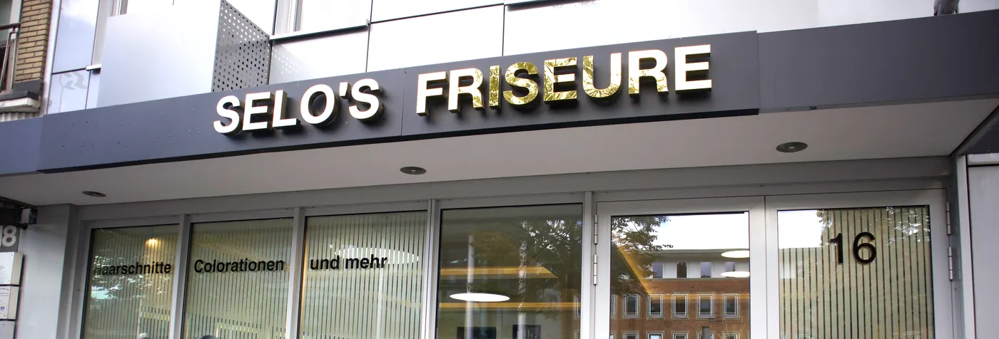 Ladenfront von SELO'S Friseure mit goldenem Schriftzug über den Schaufenstern und der Tür mit der Hausnummer 16